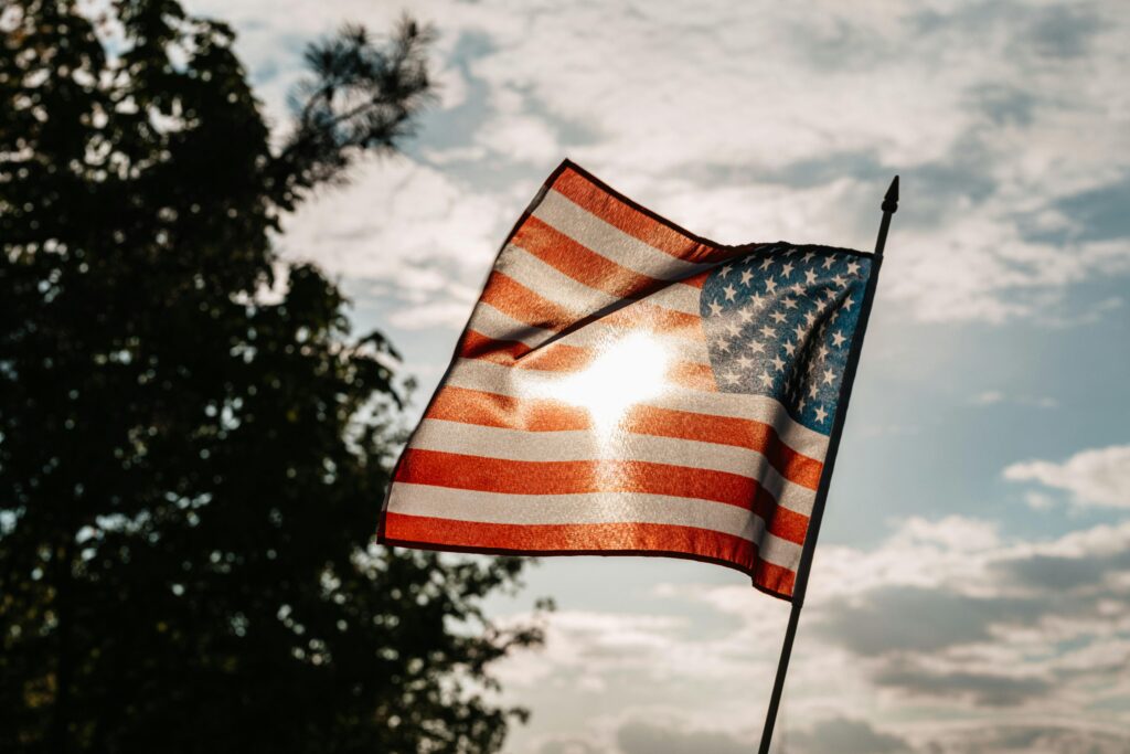 A waving American flag with sunlight streaming through against a backdrop of trees and cloudy sky.