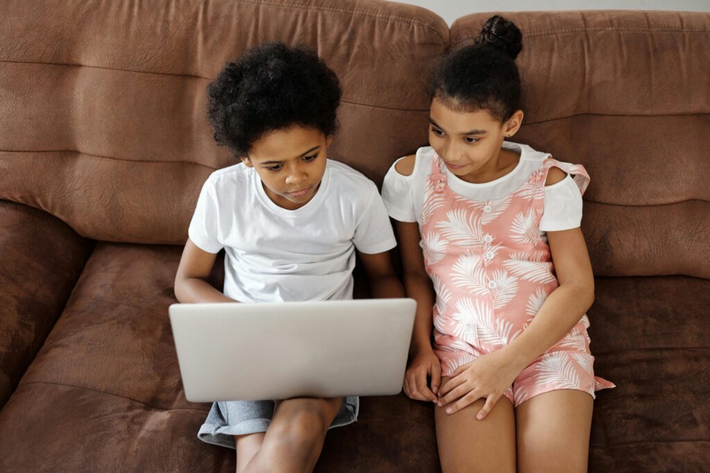 Two siblings sitting on a sofa, watching content on a laptop together indoors.