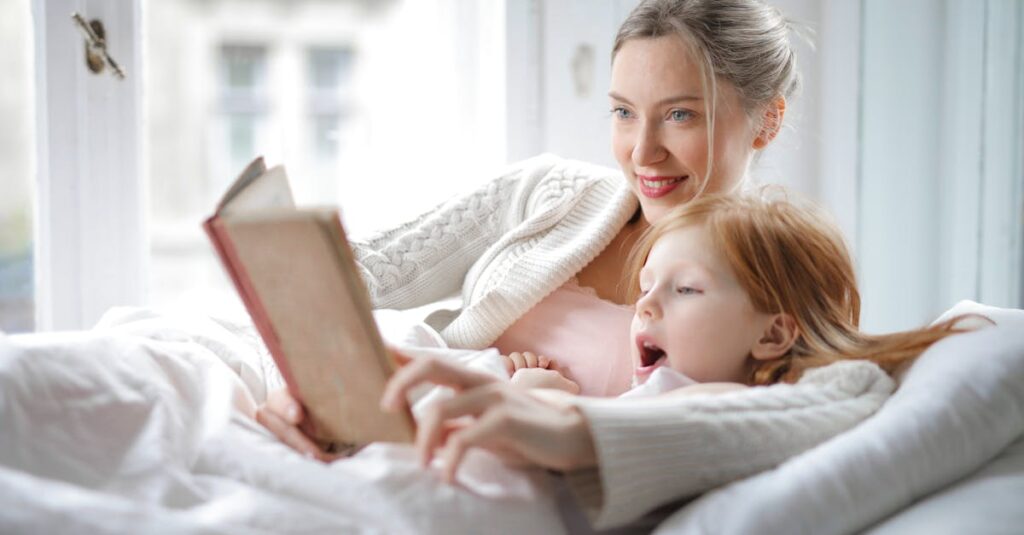 Cheerful young woman hugging cute little girl and reading book together while lying in soft bed in light bedroom at home in daytime