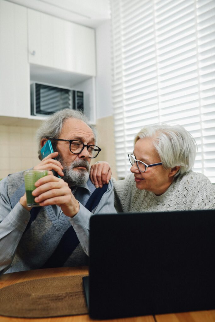 Elderly couple engaged with laptop and phone in a cozy kitchen environment.