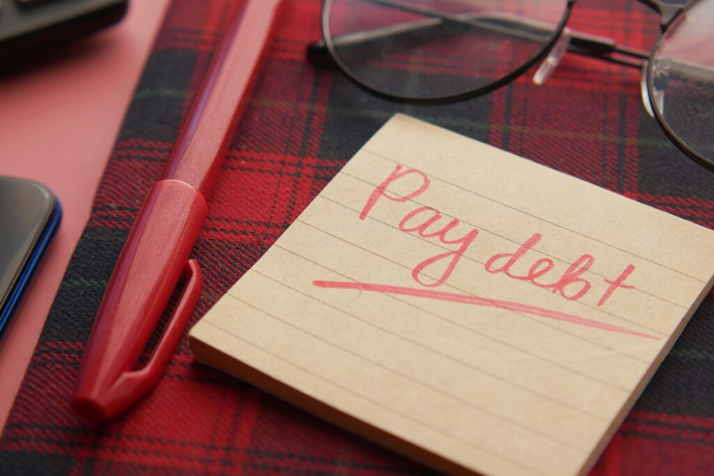 Close-up of a note reading 'Pay debt' next to a red pen on a plaid fabric, emphasizing financial reminders.